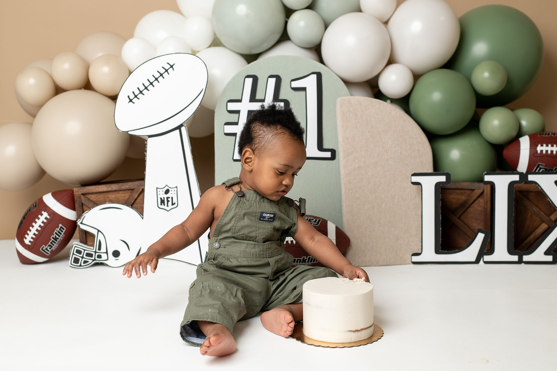 One year old touches cake during cake smash photoshoot, green balloons and football themed props in the background
