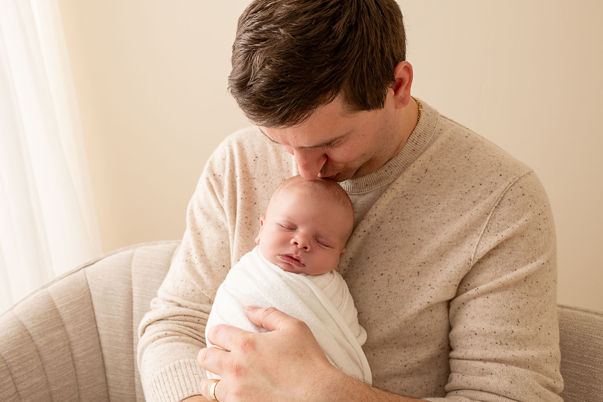 Dad kisses babies head during newborn photoshoot. tan background and couch.