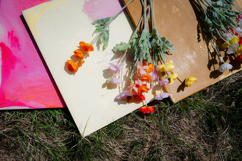 mehrere abstrakte Leinwände in Rosa-, Creme- und Sandtönen liegen im Gras mit bunten Blumen im Sonnenlicht.