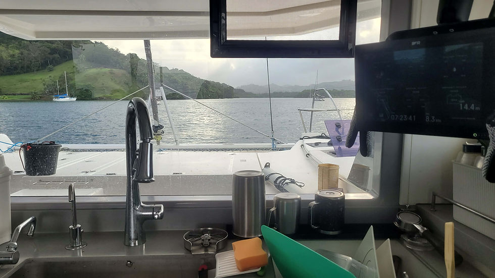 View from a boat's kitchen, showing sink and countertops in the foreground. Ocean and green hills visible through the window. Calm mood.