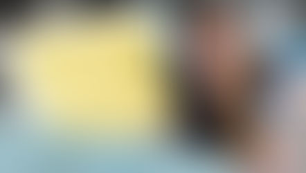 Smiling woman holds a blue electric tool and yellow fabric in a workshop. She's surrounded by folded fabric and a measuring tape around her neck.