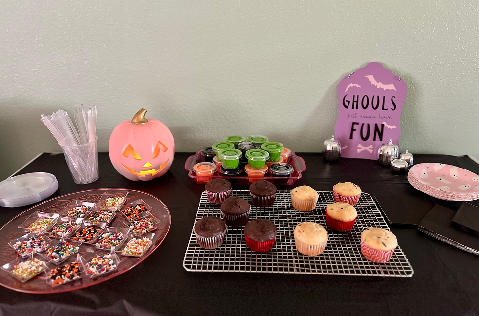 A cupcake decorating table with sprinkles, cupcakes, and frosting on it. In the background are various pumpkin and. Halloween decorations.