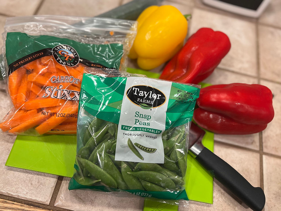 Snap peas, carrot sticks, red and yellow bell peppers, and an English cucumber on a green cutting board with a knife, on a tile counter.  
