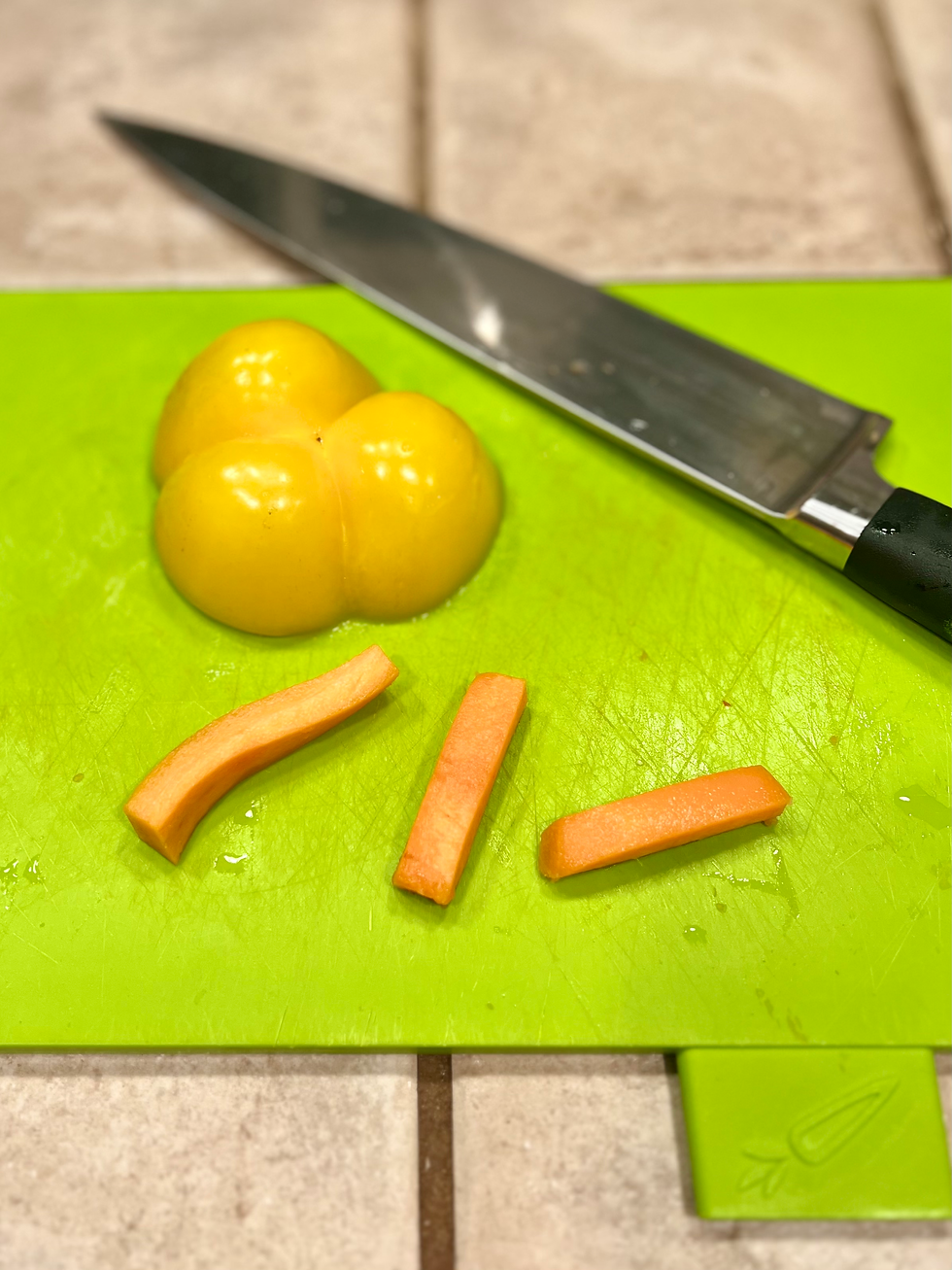 A yellow bell pepper, three carrot sticks, and a chefs knife on tops of a green cutting board on a tile counter.
