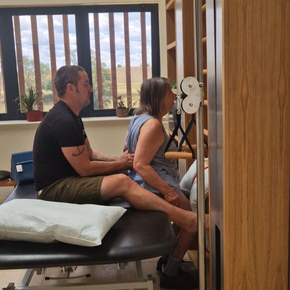 Man assists woman in a physical therapy exercise in a room with large windows and plants. Calm atmosphere.