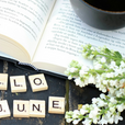 An open book with a coffee cup and a flowering branch sitting atop it. Scrabble pieces spell out "Hello June" beside. 