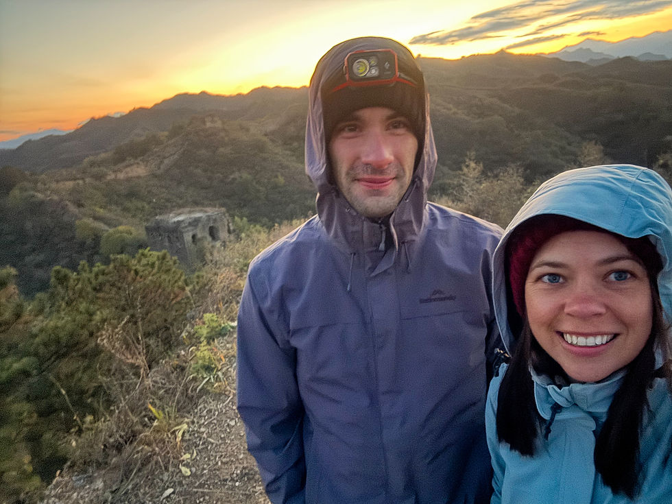 Alli and Isaac in hooded raincoats smiling at sunrise on a mountain trail, with a scenic view of hills and the Great Wall of China in the background.