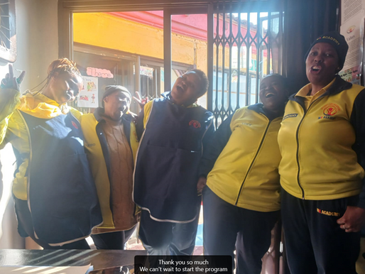 Group of smiling women in yellow and blue uniforms inside a classroom, celebrating the start of a community education programme.