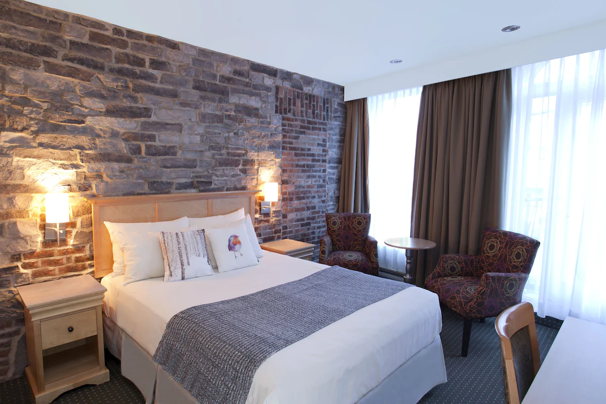 Guest room at Hôtel du Vieux‑Québec in Old Quebec City featuring exposed stone walls, warm contemporary décor, plush bedding, and modern amenities in a boutique setting.