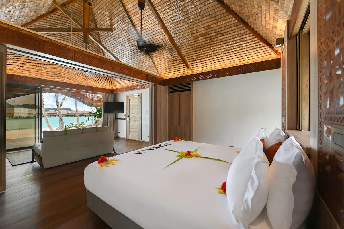 Interior of a Le Bora Bora by Pearl Resorts room featuring Polynesian‑inspired design, a bed with crisp white linens, and views of the turquoise lagoon through wide glass doors.