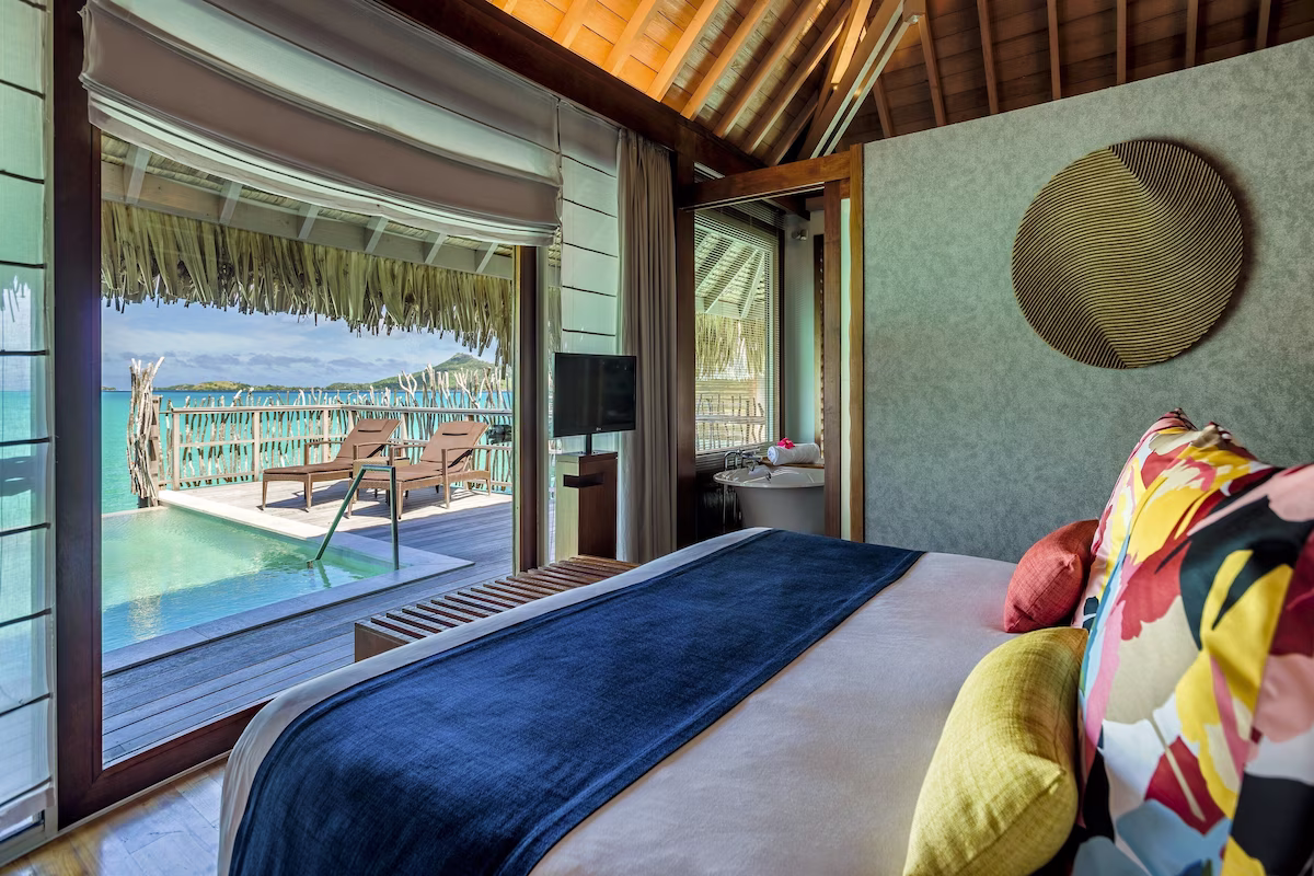 nterior of an overwater bungalow at InterContinental Le Moana Bora Bora Resort, featuring Polynesian‑inspired décor, a king bed with crisp linens, and glass doors opening to turquoise lagoon views.