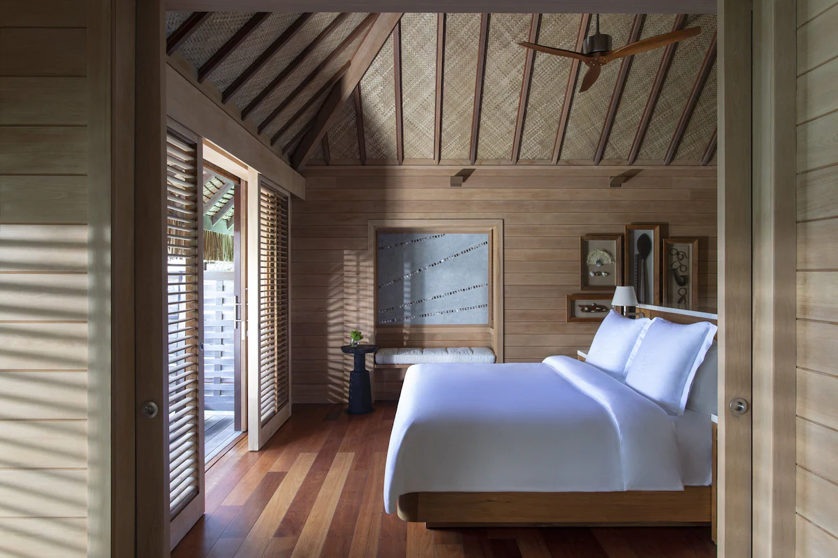 Four Seasons Resort Bora Bora overwater bungalow suite with Polynesian‑inspired décor, a canopy bed, and floor‑to‑ceiling windows opening to turquoise lagoon views.