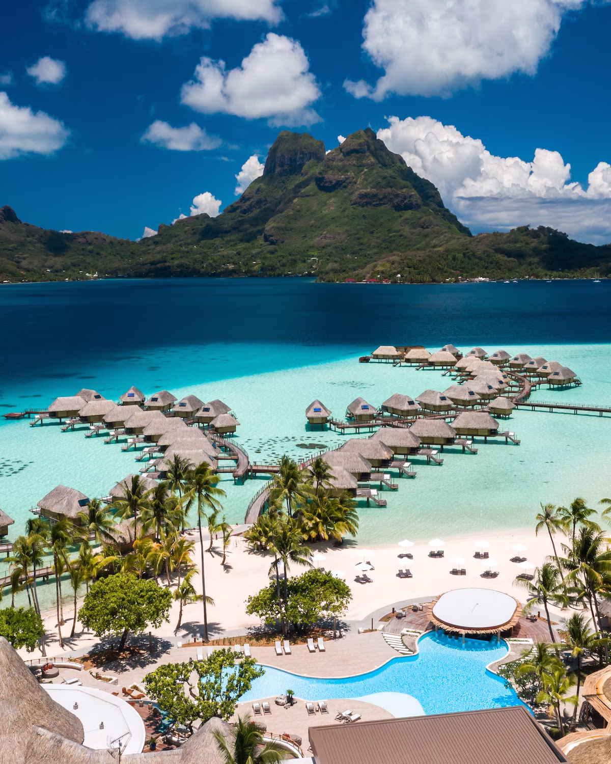 Aerial view of Le Bora Bora by Pearl Resorts, showcasing overwater bungalows perched above the turquoise lagoon with Mount Otemanu rising in the background.