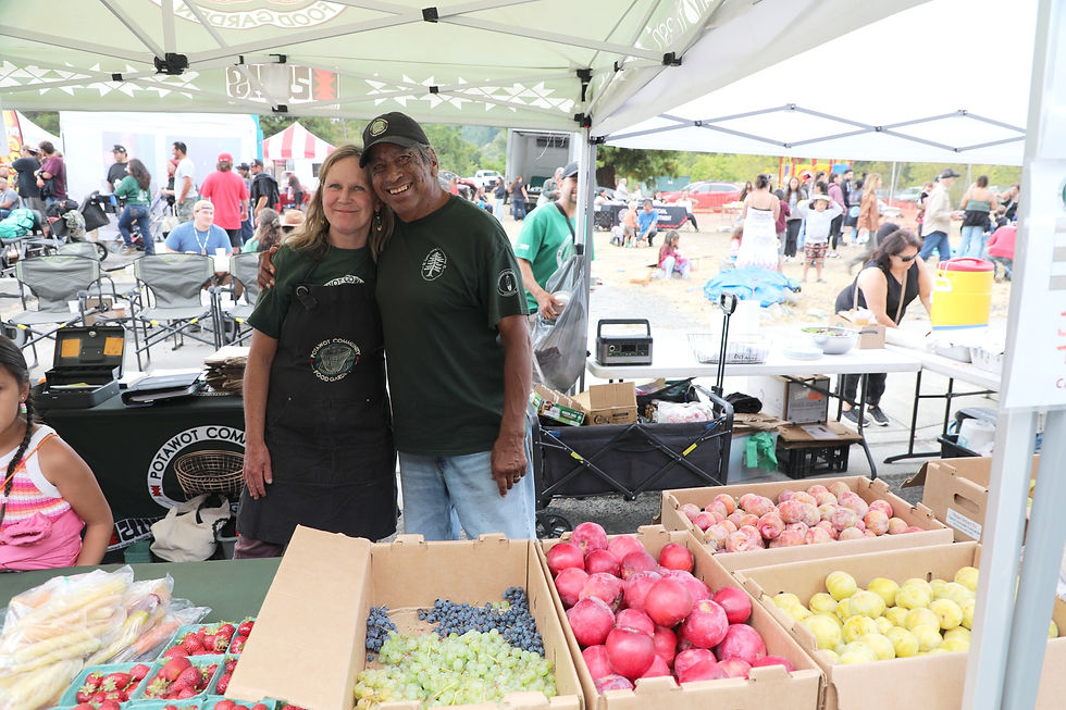Two people smiling under a green tent at a market stall with fruit boxes and "Potawot Community Food Garden" text. Busy outdoor event.