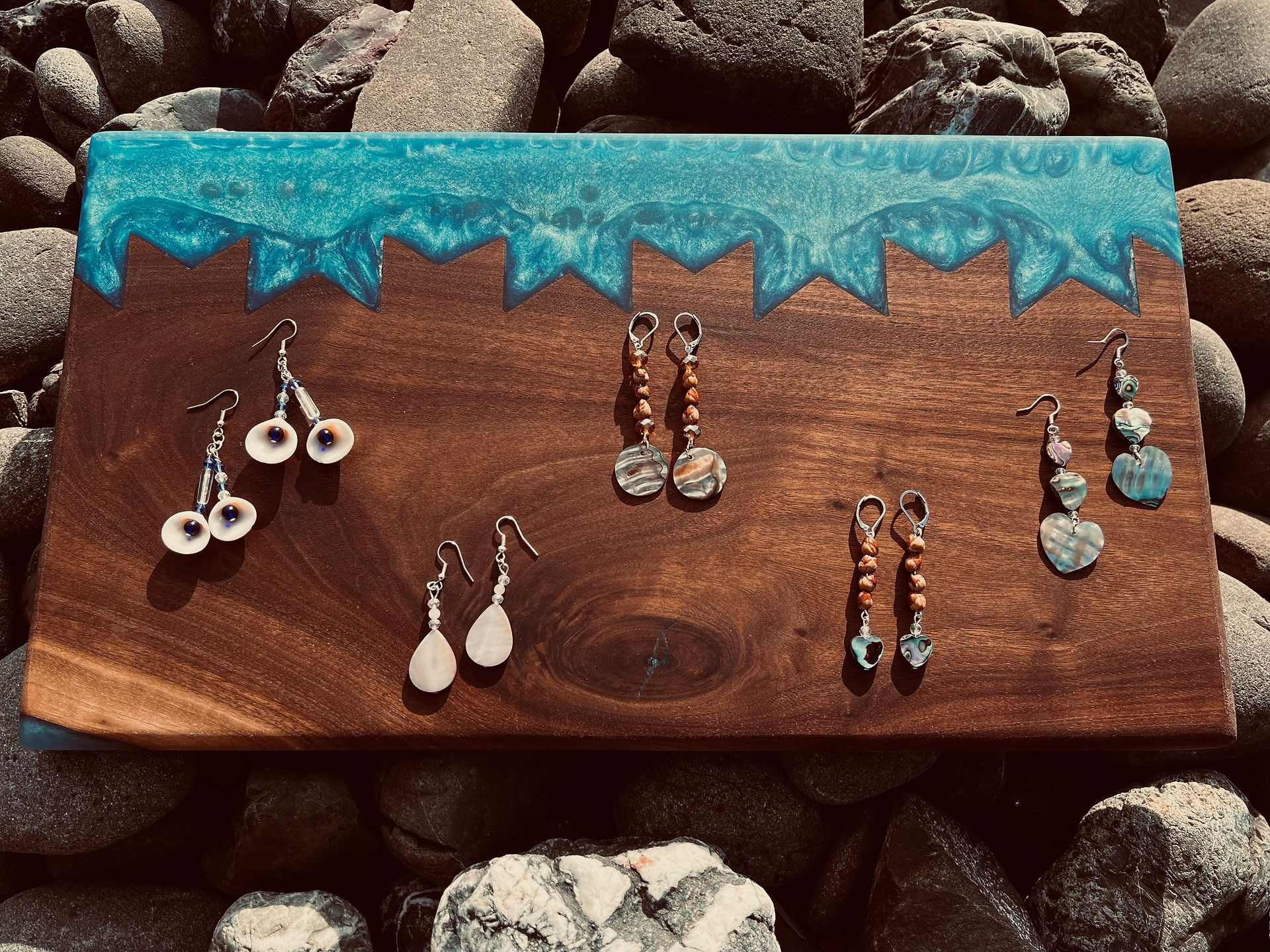 A display of various handmade earrings set on a wood plank with a blue resin wave design, laid against a rocky beach backdrop. The earrings feature designs with pearls, shells, and beads.