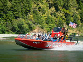People on a red "Eagle 1" jet boat waving on the Klamath River, forested background, American flag flying, text "Klamath River Jet Boat Tours."