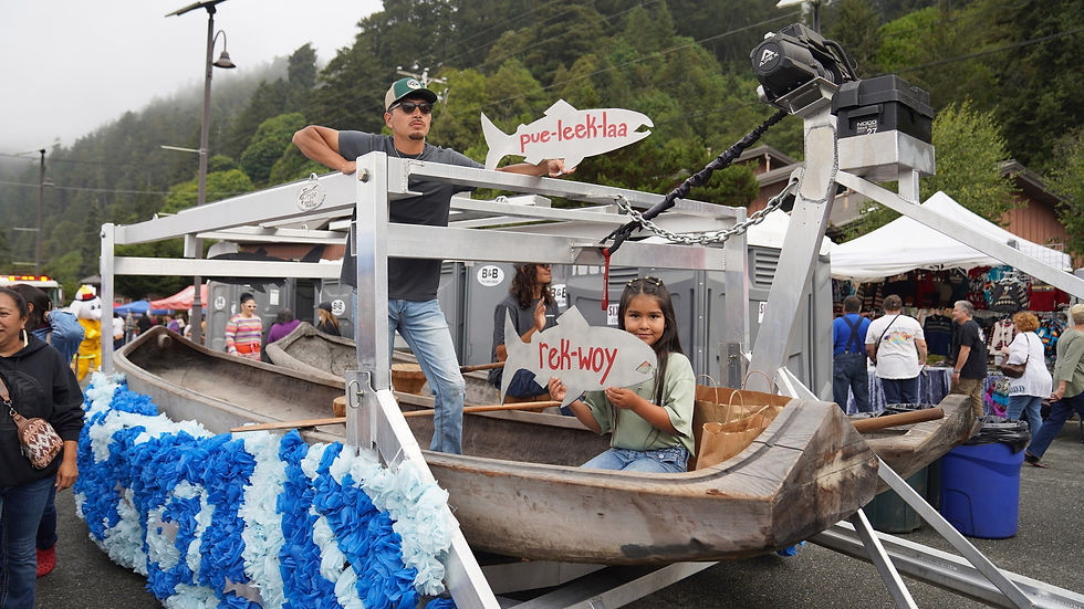 A man and girl hold fish-shaped signs in a wooden boat with blue decorations at an outdoor fair. Trees and tents are in the background.