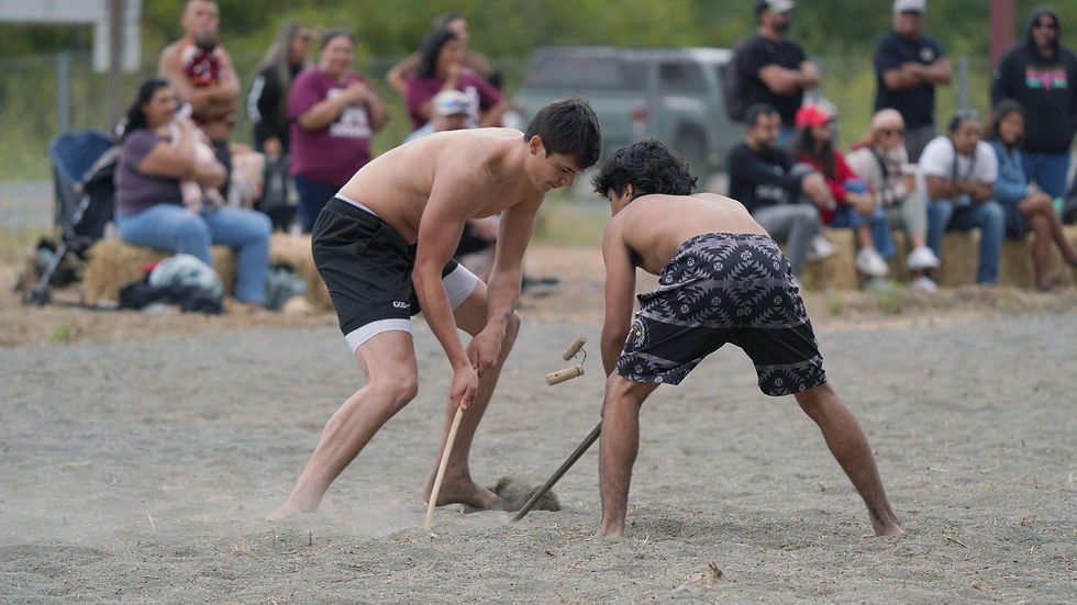 Two men, shirtless, engage in a traditional Yurok stick game on dirt. Spectators sit on hay bales watching intently. Overcast sky, casual setting.