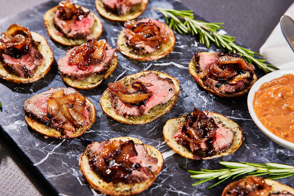 Dark marble platter displaying gluten free appetizer of herb roasted potatoes topped with seared steak, caramelized onion peppercorn jam, and roasted heirloom tomato compote, garnished with rosemary sprigs