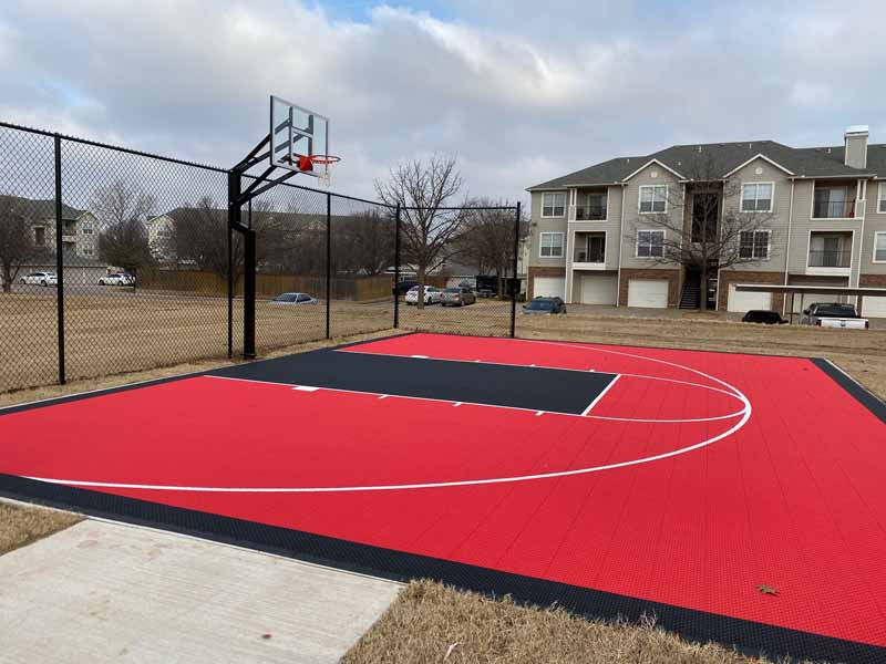 Outdoor basketball court with red and black surface, fence, and apartment buildings.