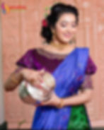 A woman dressed in traditional South Indian attire is wearing a vibrant purple and blue saree with intricate embroidery. Her