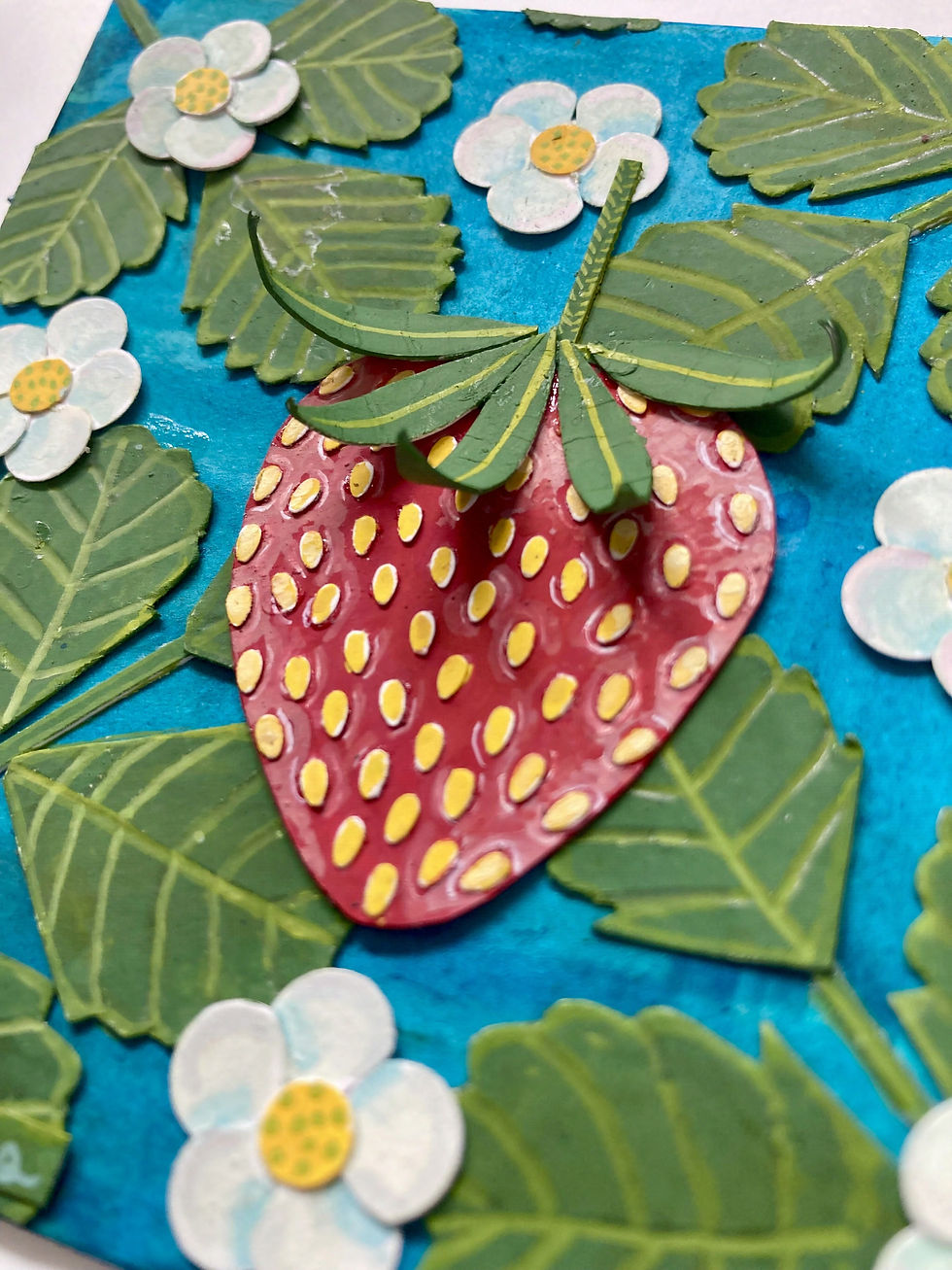 Closeup of deep teal/blue background with green strawberry leaves and white strawberry flowers with yellow centers behind one large red strawberry with yellow seeds.