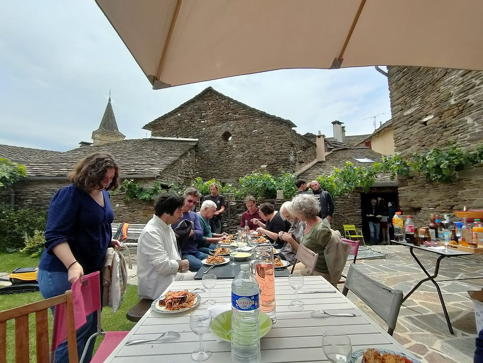 Longue table dressée dans une cour en pierre, entourée de participants, avec les toitures et le clocher du village en arrière-plan.