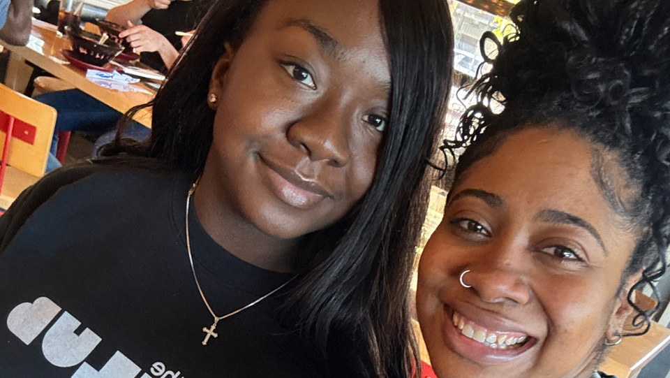 Young woman with dark skin and straight hair with another young lady with medium skin tones and her hair pulled up.