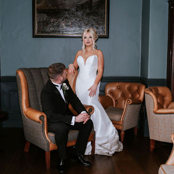 A seated groom looks towards his bride who is standing next to him in the reception room at Auchen Castle