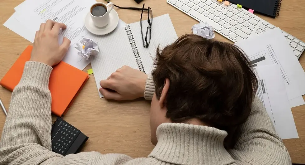 An exhausted man resting his head face down on a desk covered in paperwork