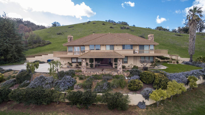 Large beige house with brown roof, stone entryway, nestled on green hillside.