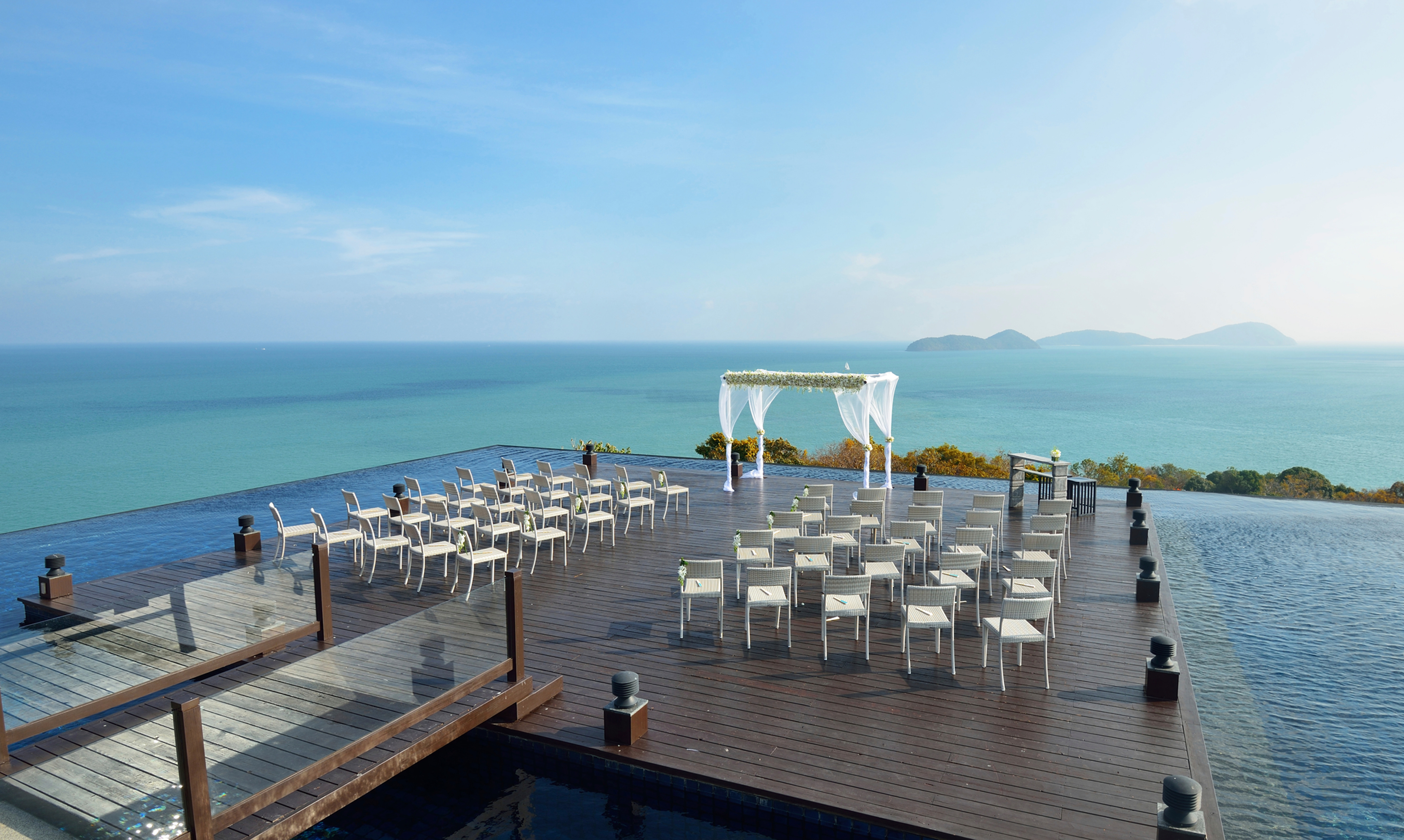 Seaside wedding deck with white chairs and floral arch overlooking a calm blue ocean and distant islands under a bright sky