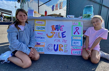Two girls sitting in front of a table  that has a poster in the front that says BAKE SALE for the Cure. 