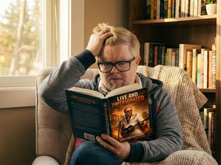 Jonathan with worried look, reading his novel, Live and Impersonated in a chair by a window with a book shelf in the background.