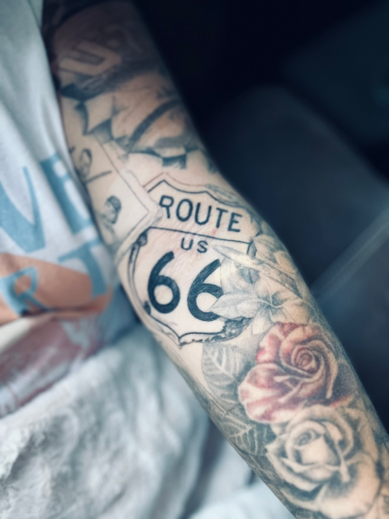 Custom sleeve tattoo in black and gray with pops of color, featuring symbolic imagery including the Route 66 highway sign, roses, vintage photographs, antlers, and a checkered racing flag. This realism-style tattoo tells a deeply personal story, highlighting meaningful moments from the client’s life through detailed, illustrative artwork.