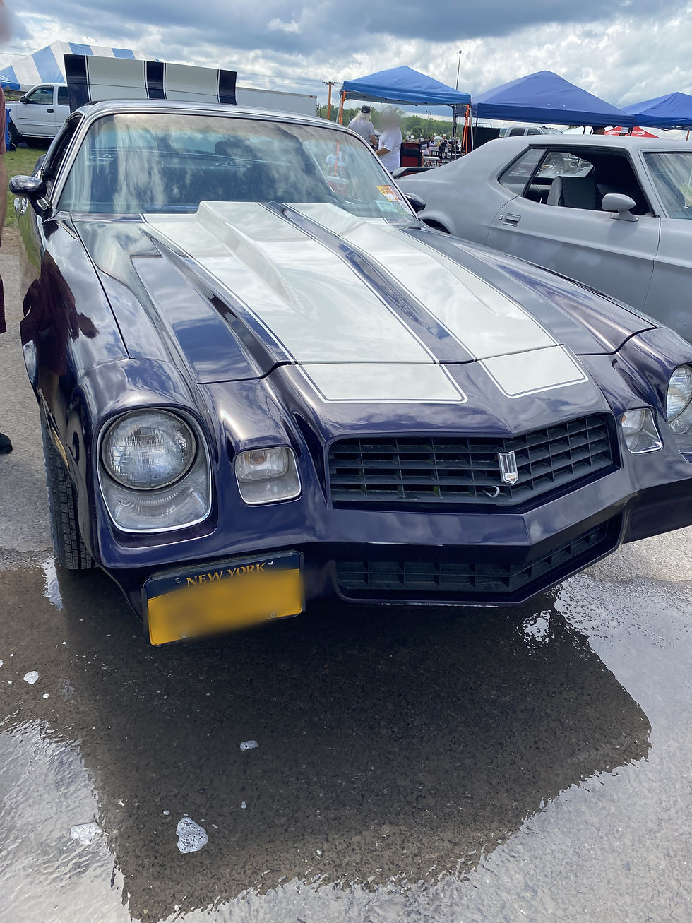 A 1980s Camaro from the front. It is blue with white racing stripes and the trunk is open.