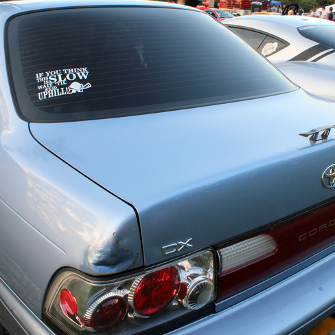 The back of a 90s Corolla, with a sticker that reads "If you think this is slow, wait til we go uphill" with a turtle next to it. It also has a mini spoiler on the back.