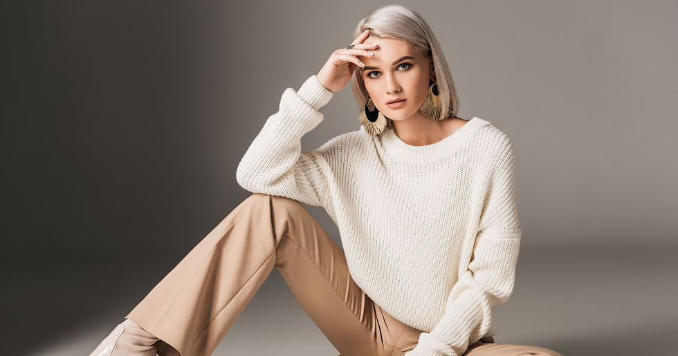 Model in an oversized cream knit sweater, beige trousers and gold statement earrings seated against a grey studio backdrop.