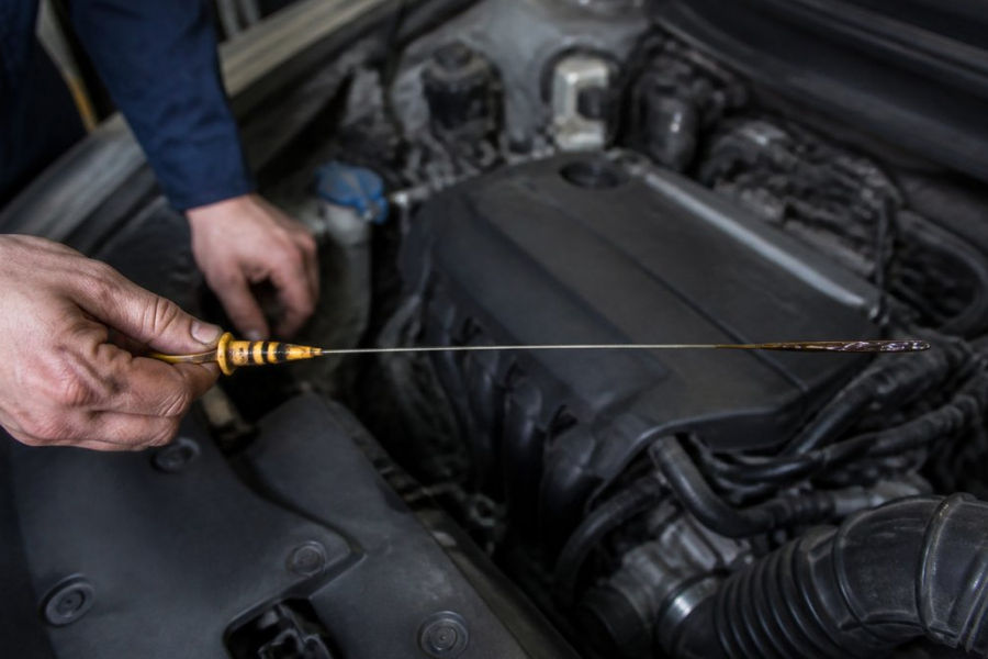 Mechanic's hands pulling out an oil dipstick from under a car hood, showing the dipstick fully extended with dark oil residue visible on the yellow handle and tip