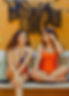 two girls sitting on a bench which one with a swimwear piece. A plain orange bikini bottom with a print bikini top, and a plain swimsuit in orange made with a sustainable fabric