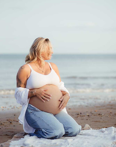 An expectant mum kneeling on a rug at the beach and looking away into the distance.