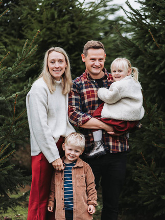A family of four smiling and laughing in front of Christmas trees.