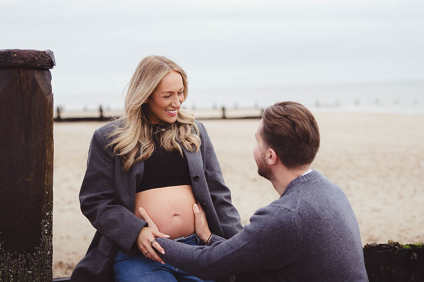 A pregnant woman sat on a wave breaker at the beach smiling at her partner kneeling and holding her baby bump.