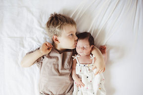 A little boy laying down cuddling his new baby sibling and kissing her on the head.
