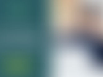 Smiling person in blue plaid shirt with headset, sitting at a desk using a laptop. Bright room with blinds.