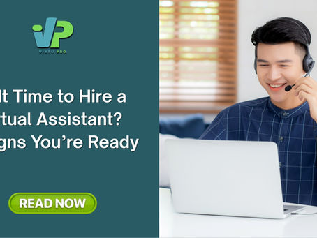 Smiling person in blue plaid shirt with headset, sitting at a desk using a laptop. Bright room with blinds.