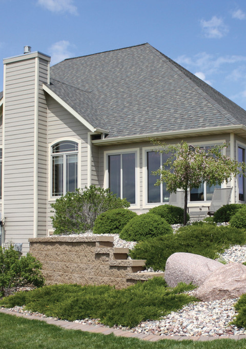 Large house with stone retaining walls, green bushes, and blue sky background.