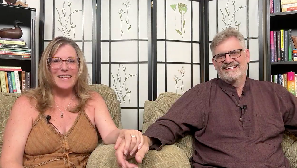 Smiling couple holding hands, seated on a couch with green cushions. Bookshelf and decorative screen with plant patterns in the background.