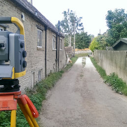 Surveying total station on tripod beside stone cottages on a narrow dirt lane, with fences and trees under an overcast sky.