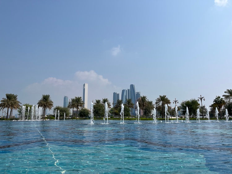 At the Presidential Palace Qasr Al Watan water fountains spray in a pool with palm trees and skyscrapers in the background under a clear blue sky in a serene, urban setting of Abu Dhabi.